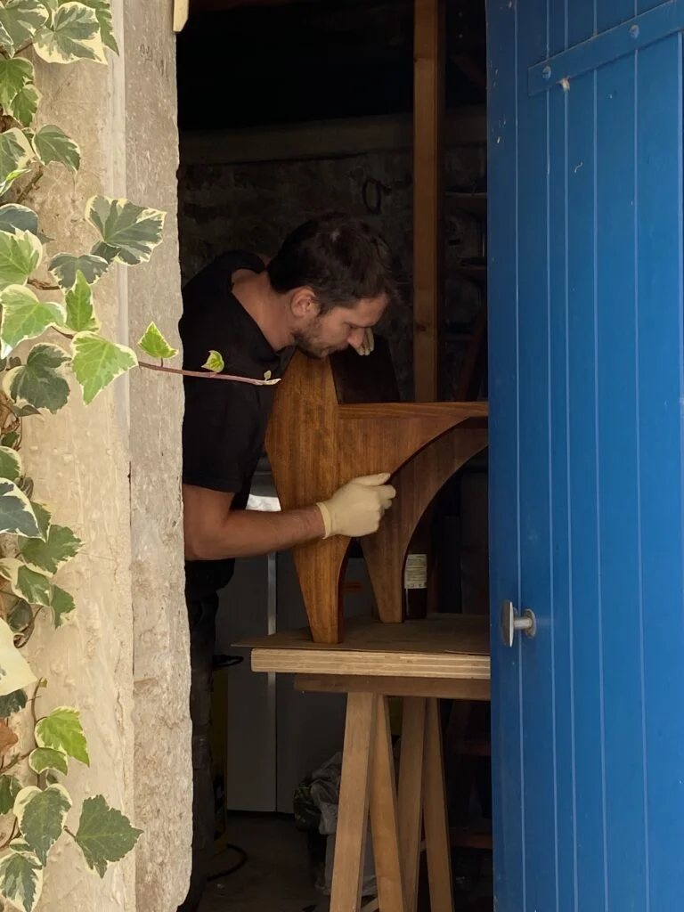 Mathieu Houlbert travail aux finitions d’un fauteuil en hêtre dans son atelier. Porte bleu ouverte.